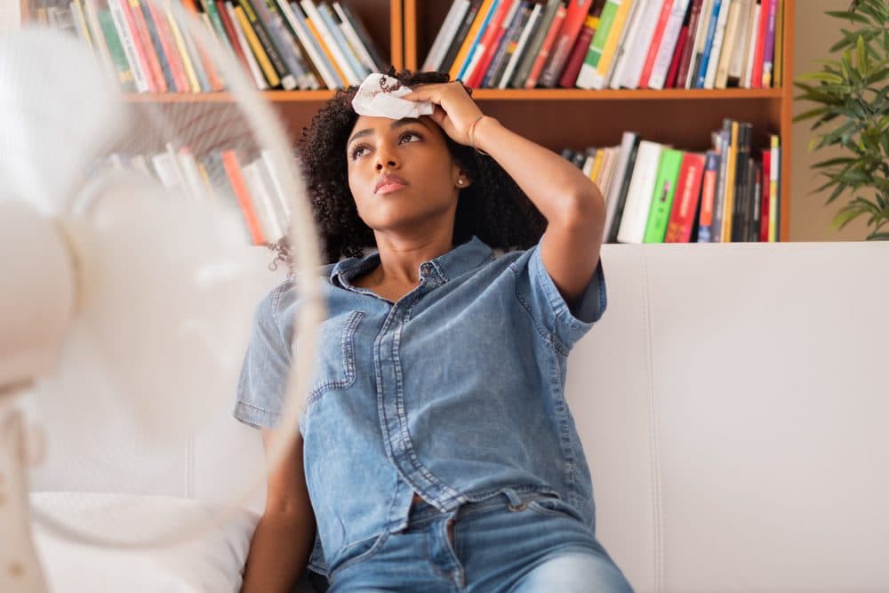 A fan is blowing on a woman who is trying to cool herself off with a damp cloth while sitting on a couch