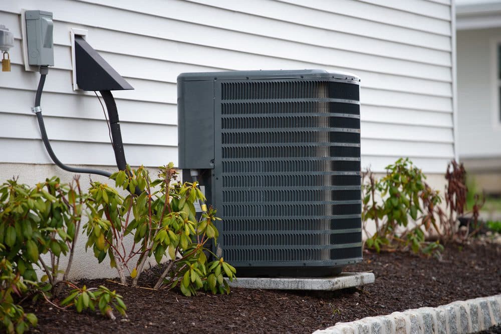 An outdoor AC unit sitting in a mulch flowerbed next to a house with white siding