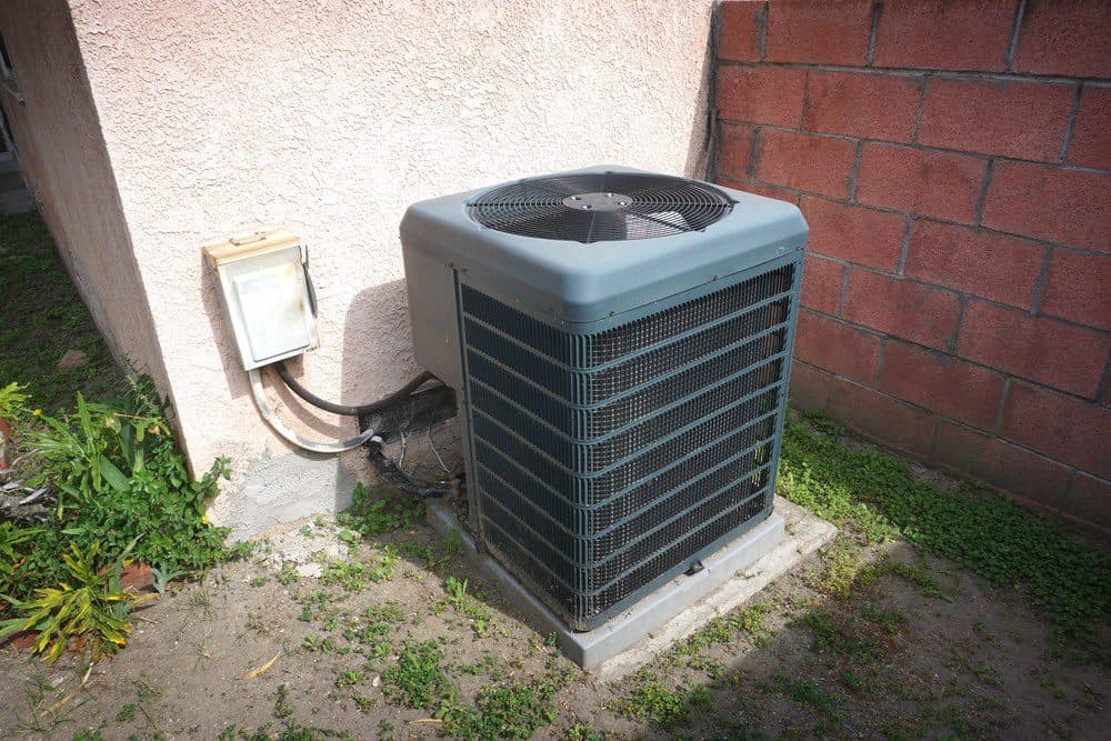An air conditioning unit connected to a house and sitting adjacent to a brick wall