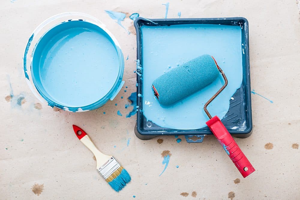 A paint brush and roller are doused in blue paint with a can full of paint next to them