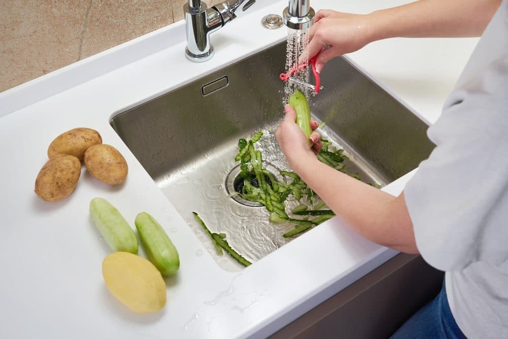 A woman peels potatoes and cucumbers in her sink with peelings left inside of the basin