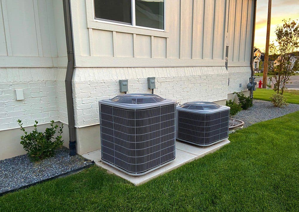 Two HVAC units outside next to a beige home with manicured landscaping