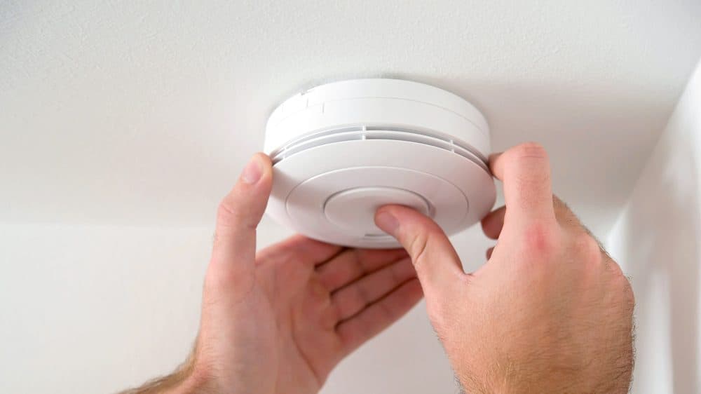 A smoke detector on the ceiling with a Casteel technician's hands reaching up to it
