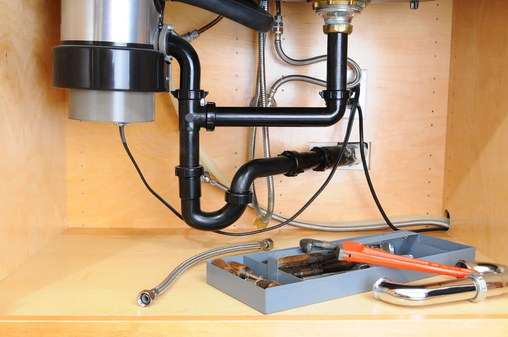 A garbage disposal is inside of a kitchen cabinet with tools to perform repairs on it are laying on the cabinet bottom