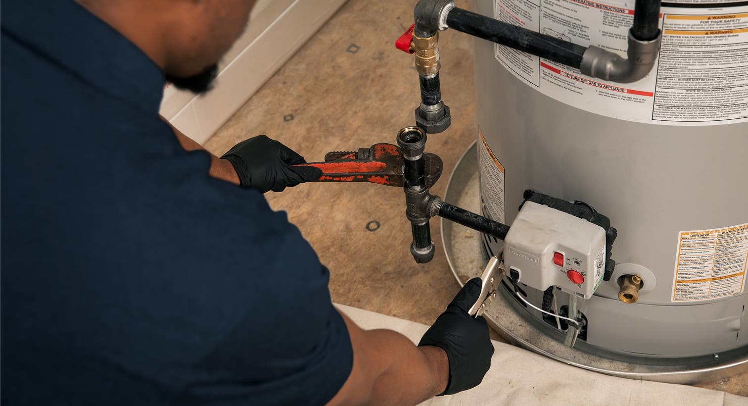 A Casteel plumber wearing black gloves uses a pipe wrench to repair the gas line and control valve assembly on a residential water heater tank.